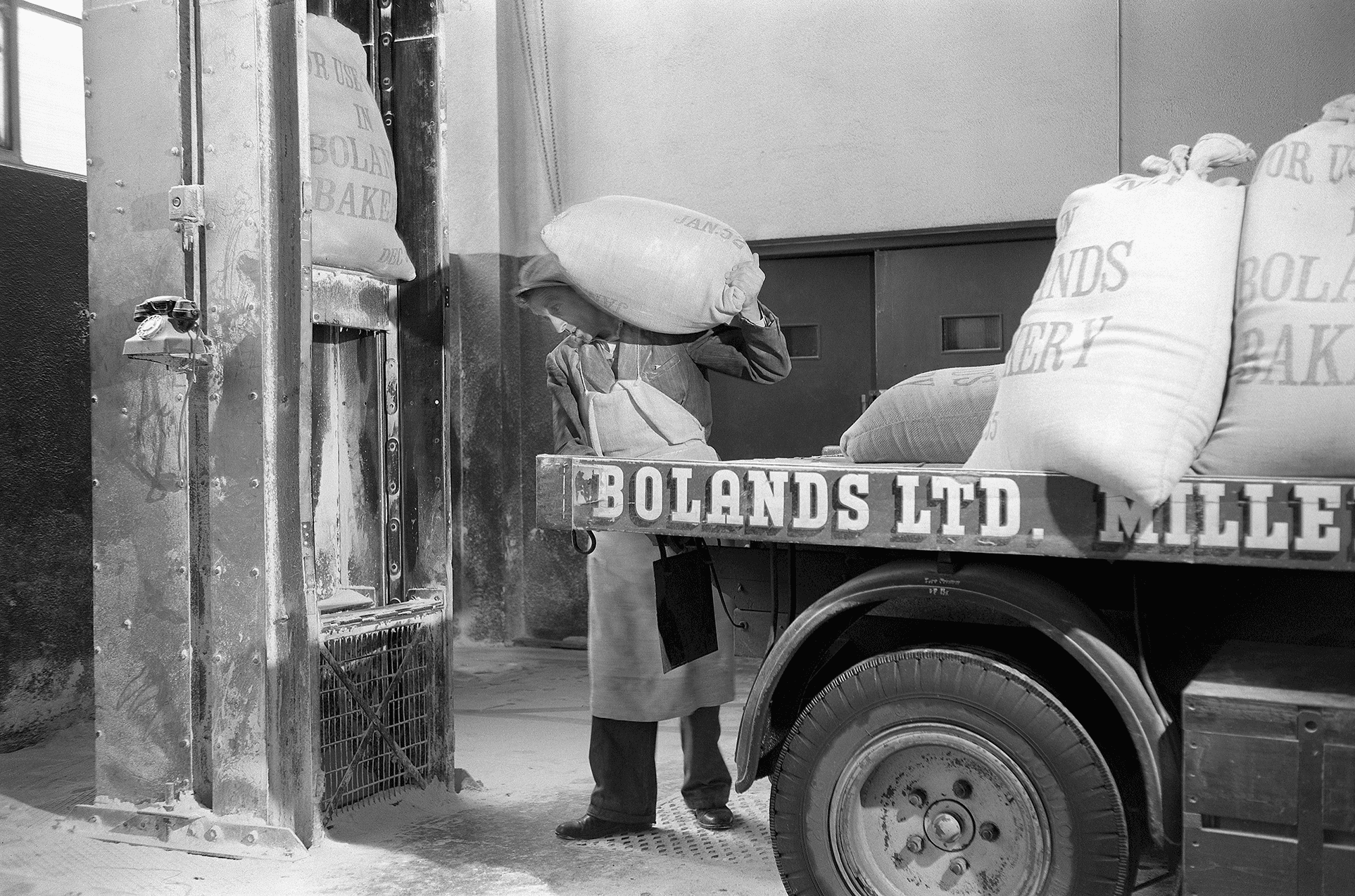 Historic black-and-white photo of a former Bolands Mills worker transferring a large sack of flour from a truck, to a freight lift on the factory floor.